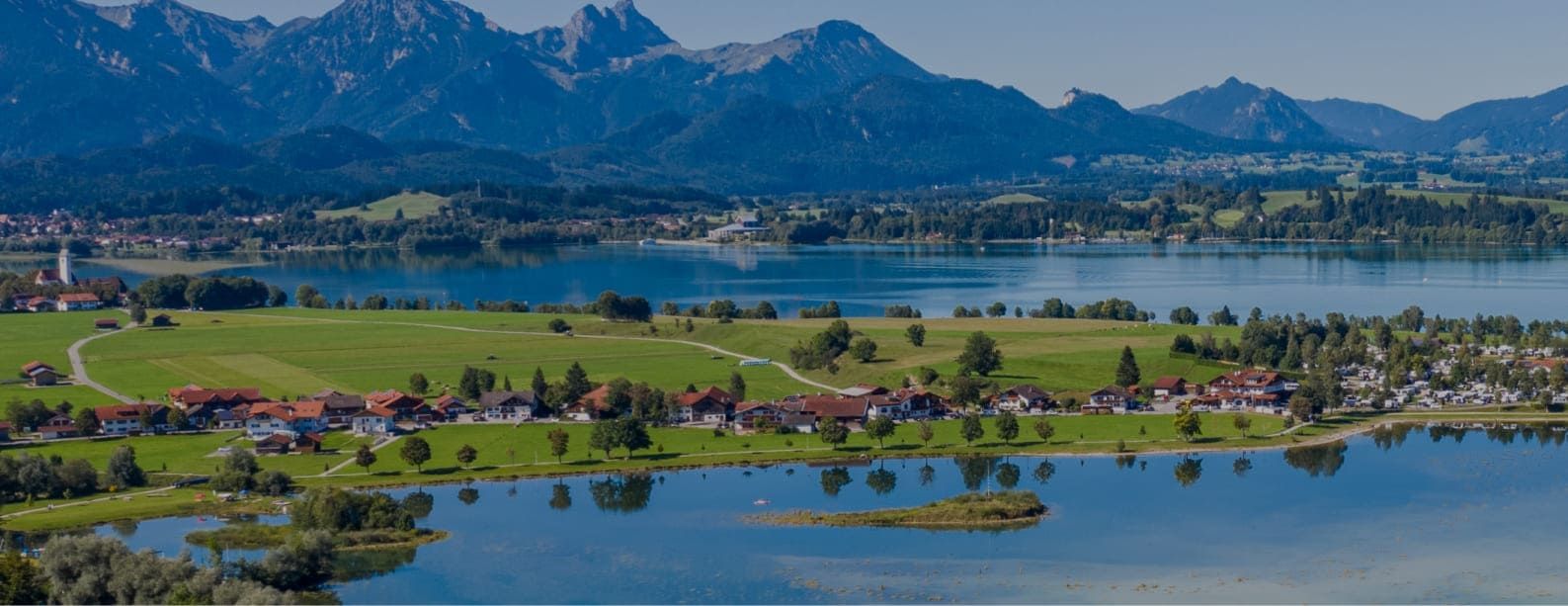 Luftaufnahme eines kleinen Dorfs am Forggensee mit grünen Feldern und kleiner Insel im Wasser